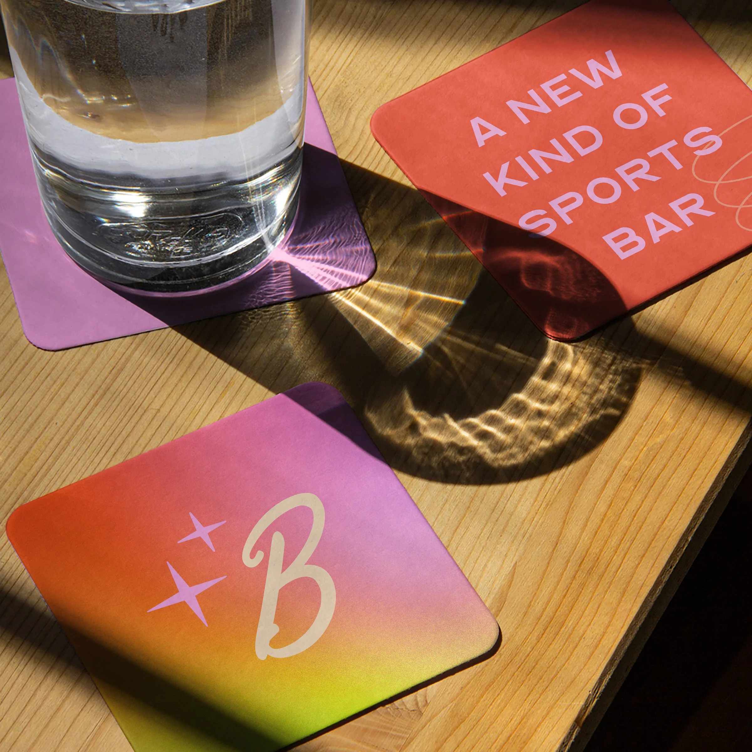 A glass of water on a purple coaster, with colorful coasters and a drink menu on a wooden table. The menu reads 'A New Kind of Sports Bar'.