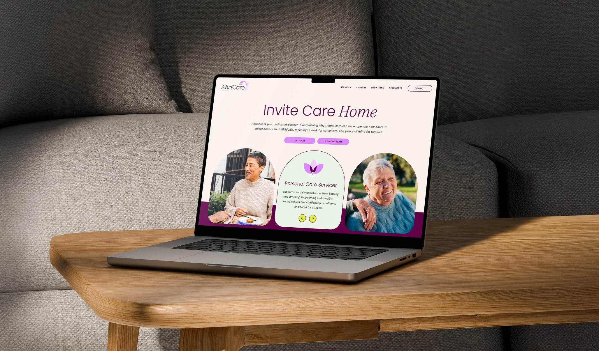 Laptop displaying a website for AbriCare's Invite Care Home services, featuring images of smiling elderly individuals and information about personal care services. The laptop is on a wooden table next to a gray cushioned sofa.