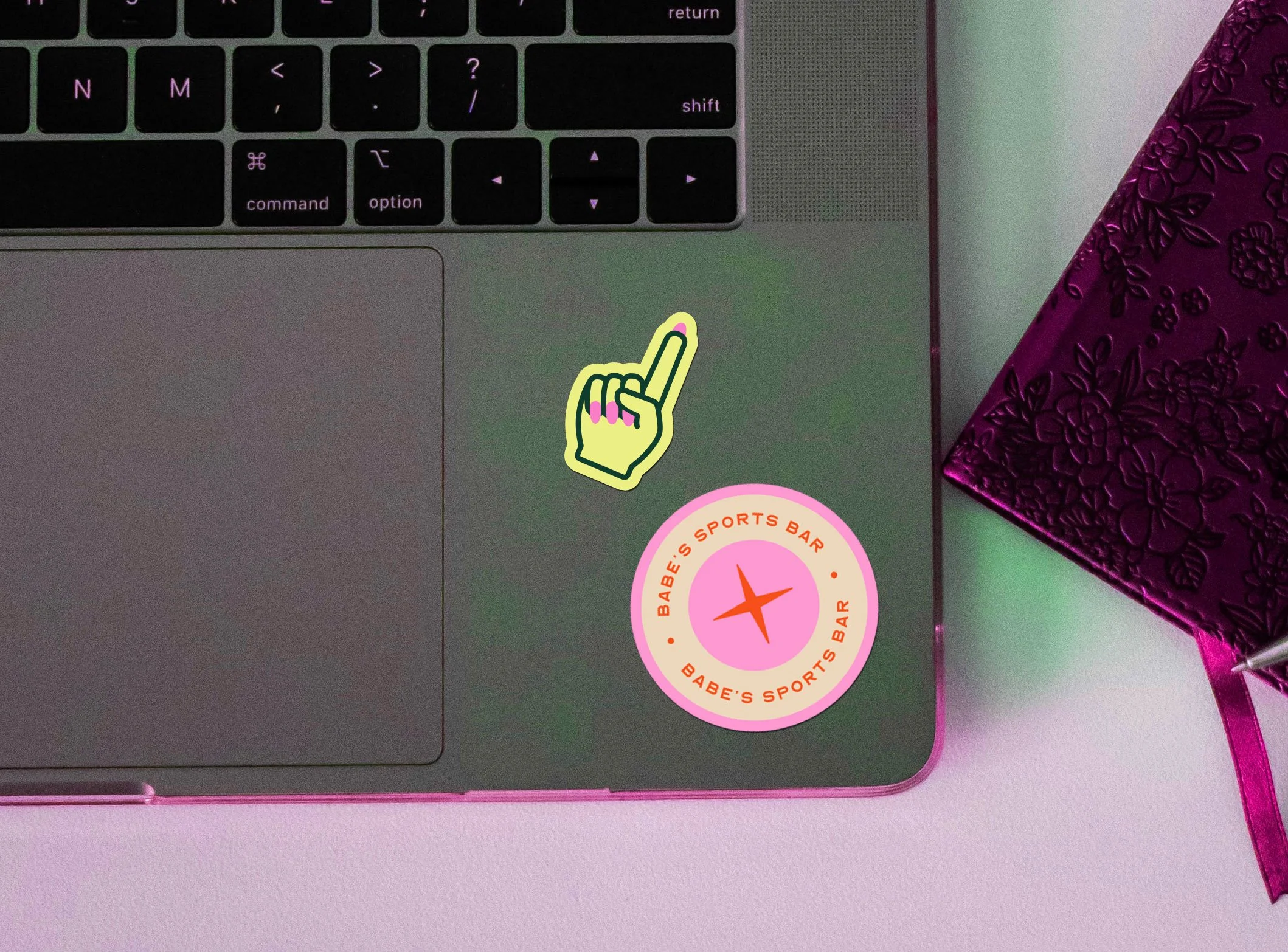 A laptop keyboard with two stickers, one showing a cartoon hand with pink nails and the other a pink and orange logo for Babe's Sports Bar, on a pink desk next to a purple floral-patterned notebook.