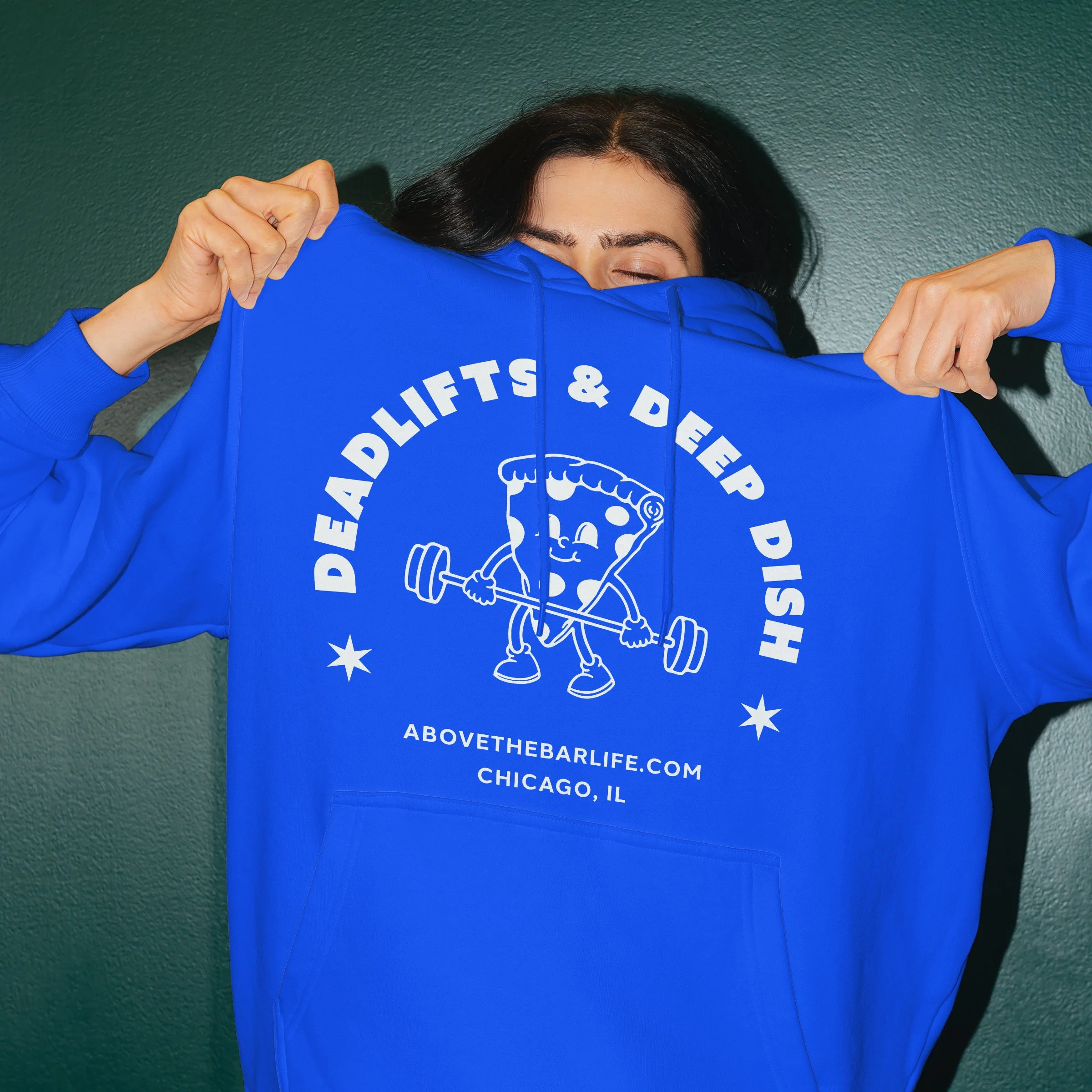 Person wearing a blue hoodie with the slogan 'Deadlifts & Dip Dishes' and a cartoon pizza lifting a barbell, standing against a green wall.