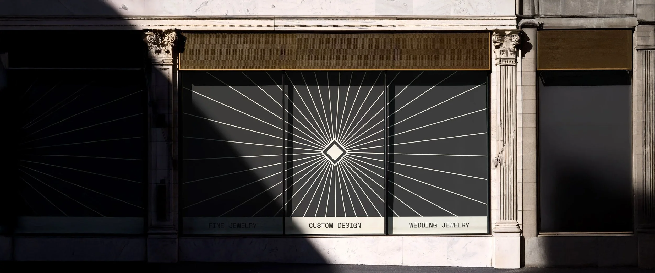 Storefront window with a black background and white geometric design radiating from a central diamond shape. Text at the bottom reads 'Fine Jewelry,' 'Custom Design,' and 'Wedding Jewelry.' Flanked by ornate stone columns.