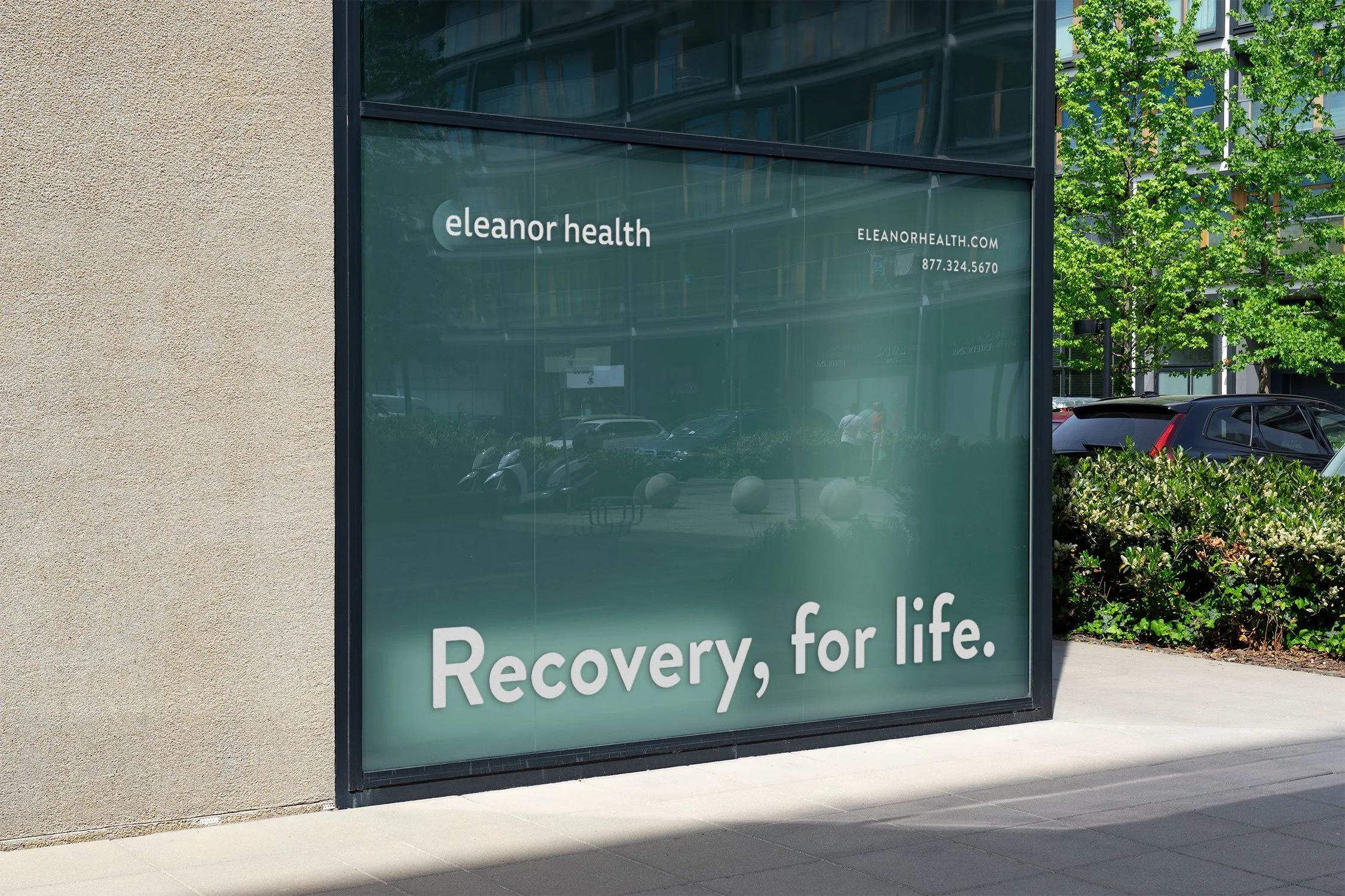 Glass window of Eleanor Health clinic with the slogan 'Recovery, for life.' and contact information, located in an urban area with trees and parked cars outside.