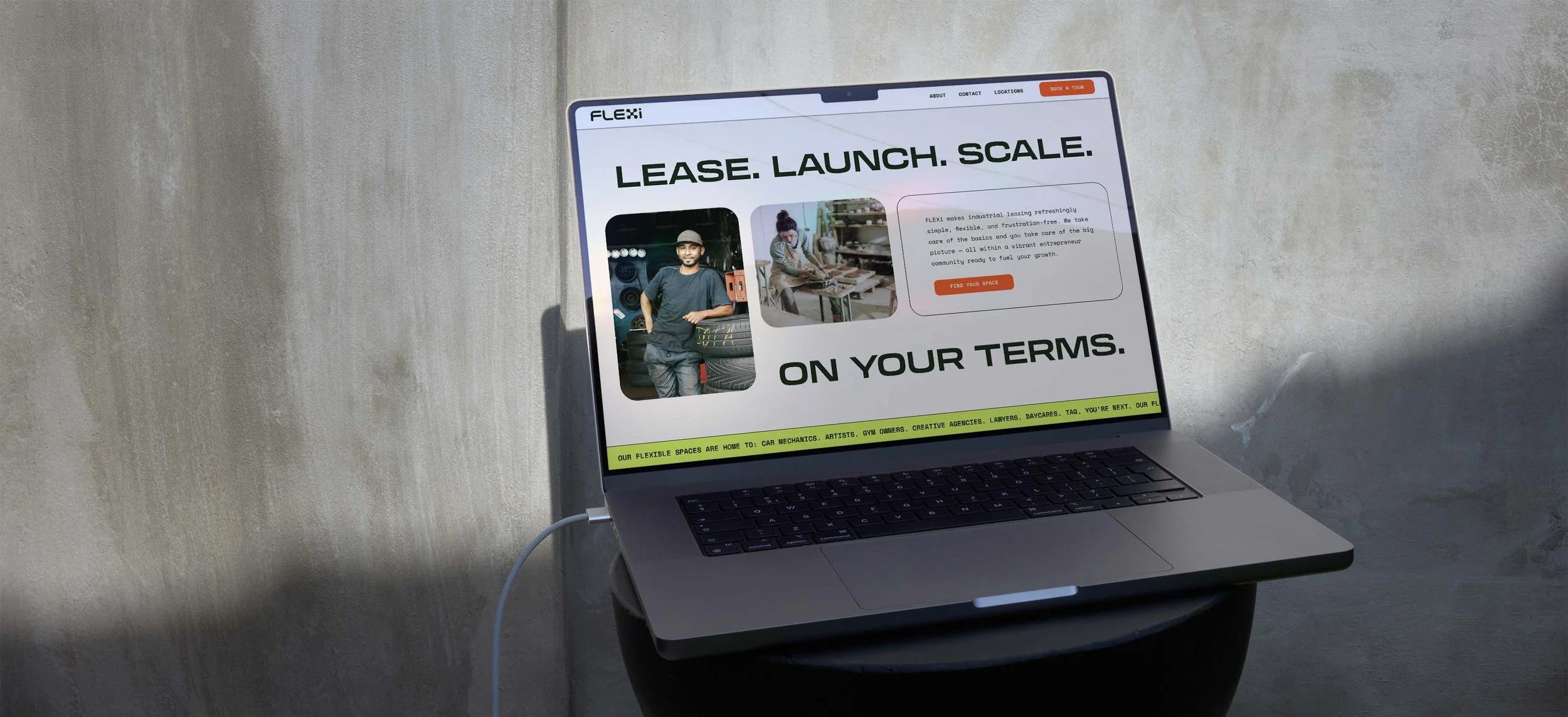 Open laptop on a stand displaying a website titled "LEASE. LAUNCH. SCALE. ON YOUR TERMS." with images of people working in a workshop and a description about flexible space leasing, against a textured gray wall background.