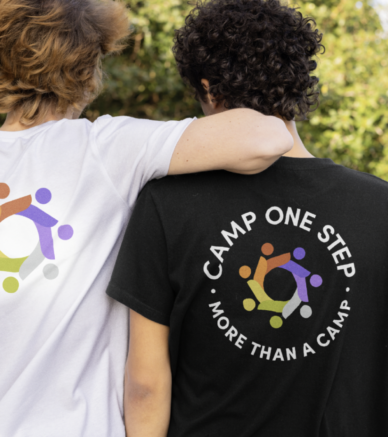 Two people wearing t-shirts with a circular logo and the slogans "Camp One Step" and "More Than a Camp". The person on the left wears a white shirt, and the person on the right wears a black shirt. They are outdoors in front of greenery, with the person on the left having their arm around the other's shoulder.
