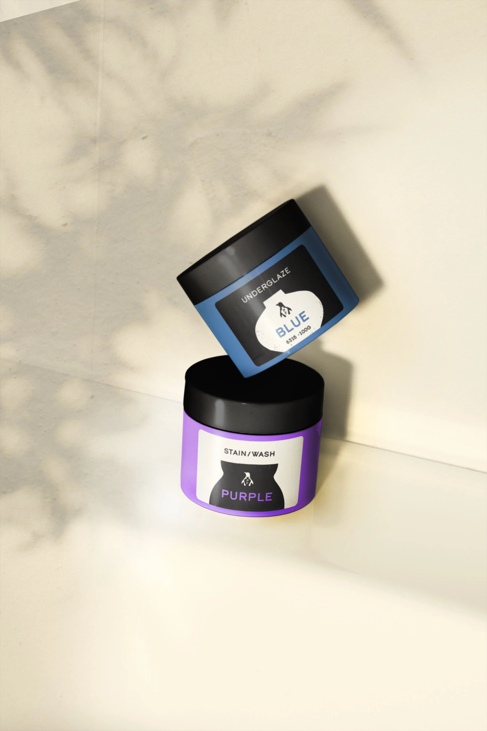 Two jars with black lids, one labeled 'underglaze blue' in blue and one labeled 'stain/wash purple' in purple, placed against a light background. The purple jar is resting on the blue jar.