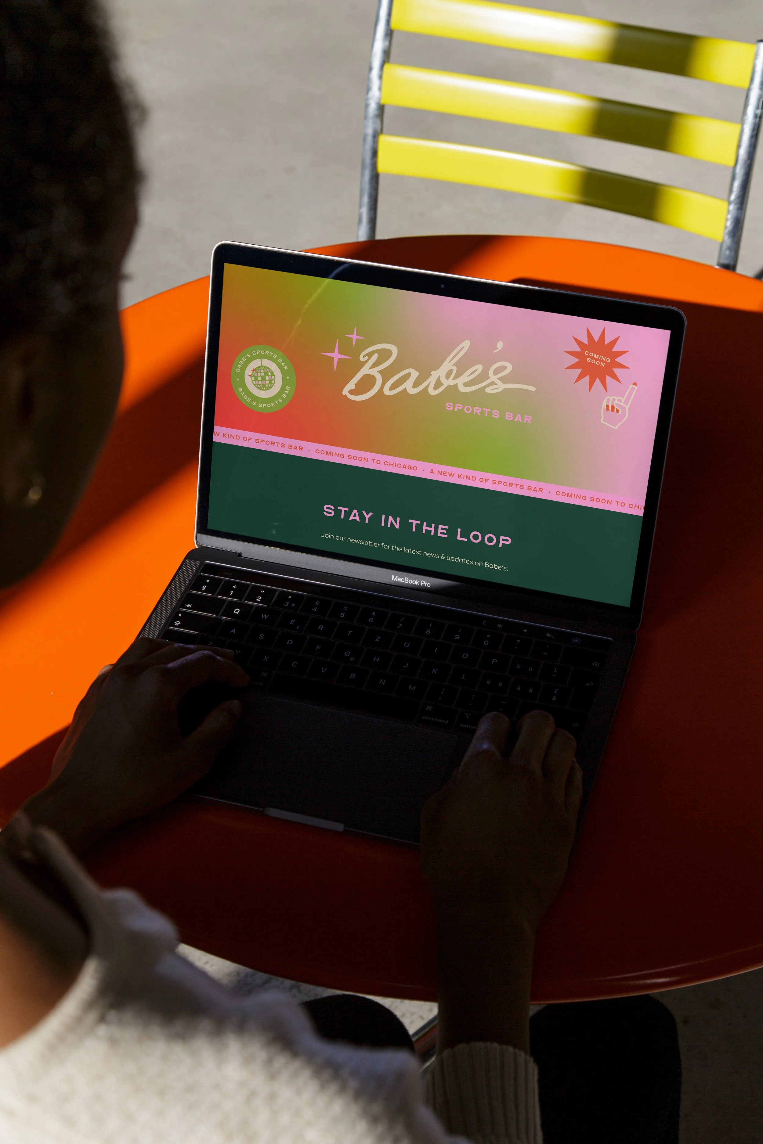 Person using a laptop at a red table displaying a website for Babe's Sports Bar with bright pink and green colors, a logo, and the message 'STAY IN THE LOOP'.
