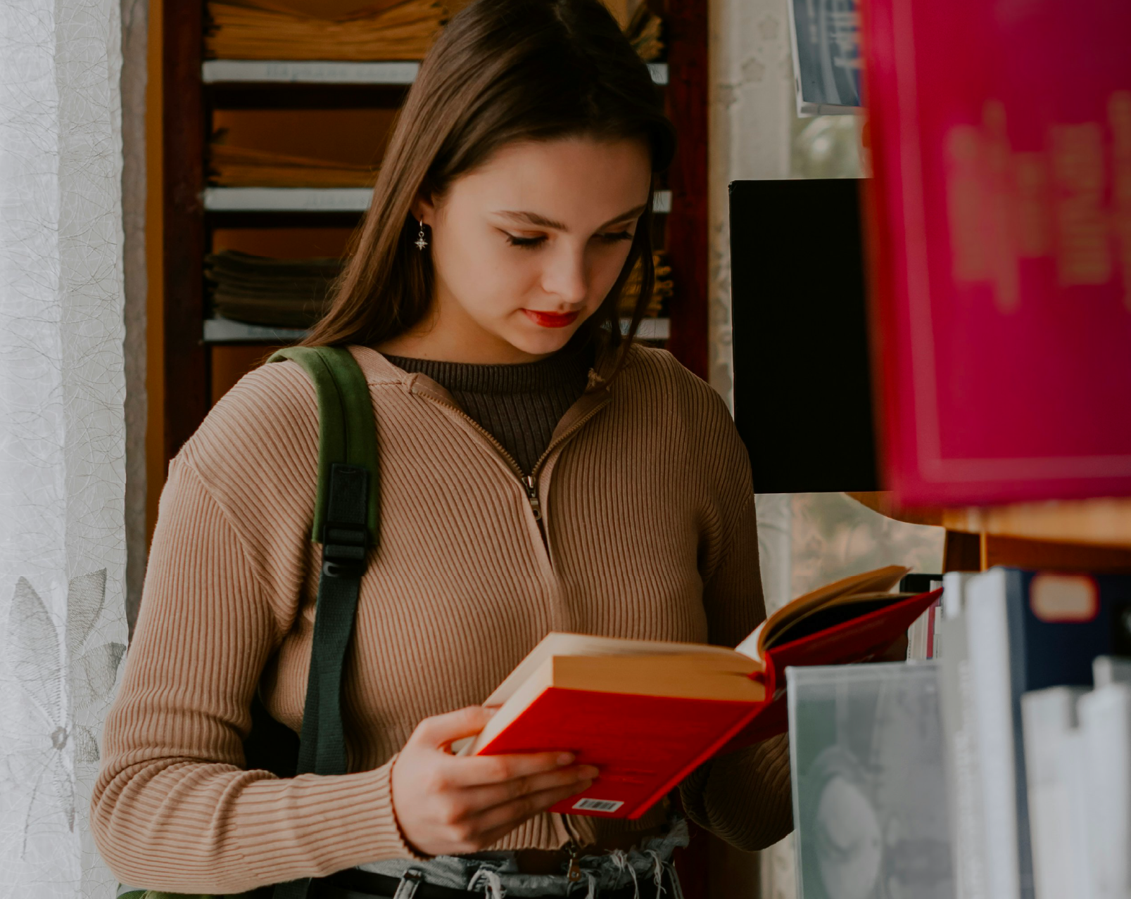 Young woman with a green strap backpack reading a red hardcover book at a bookstore.