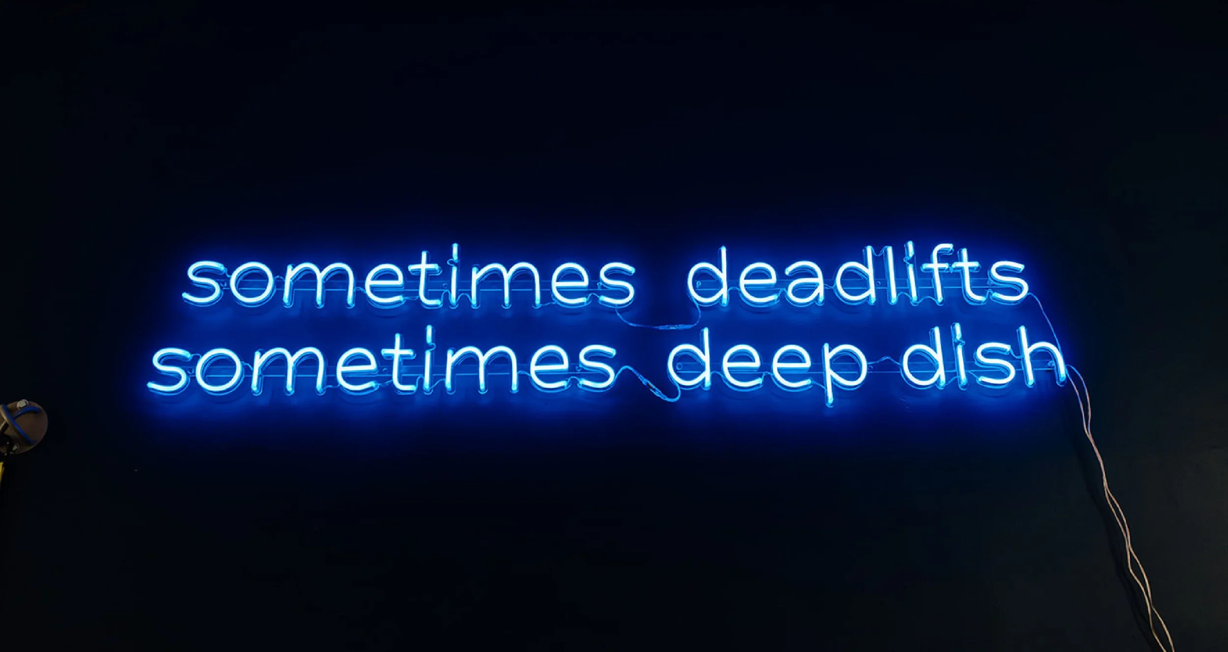 Neon sign in blue reading, "sometimes deadlifts sometimes deep dish" against a black background.