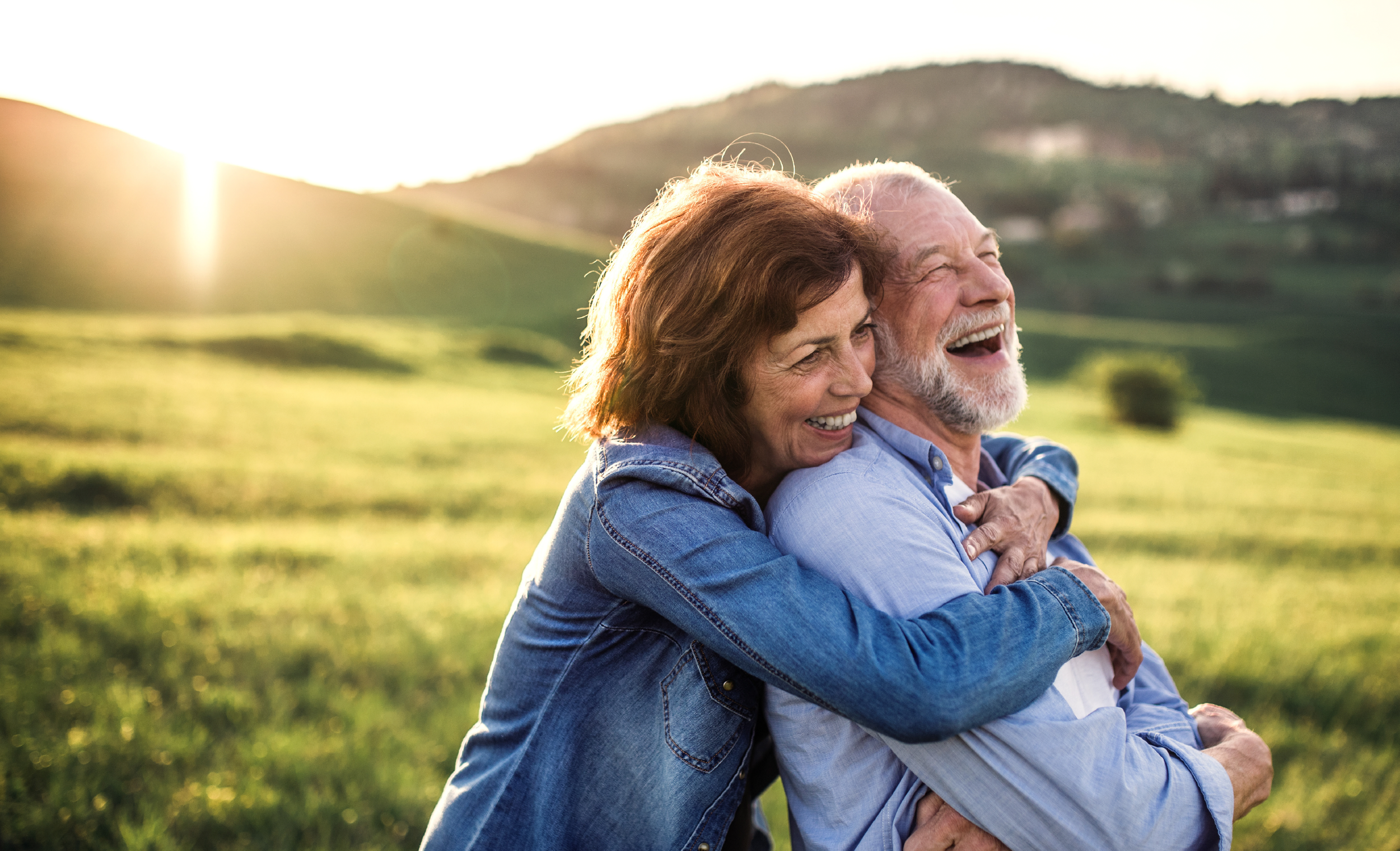 Senior couple hugging outdoors on a sunny day with green hills in the background.