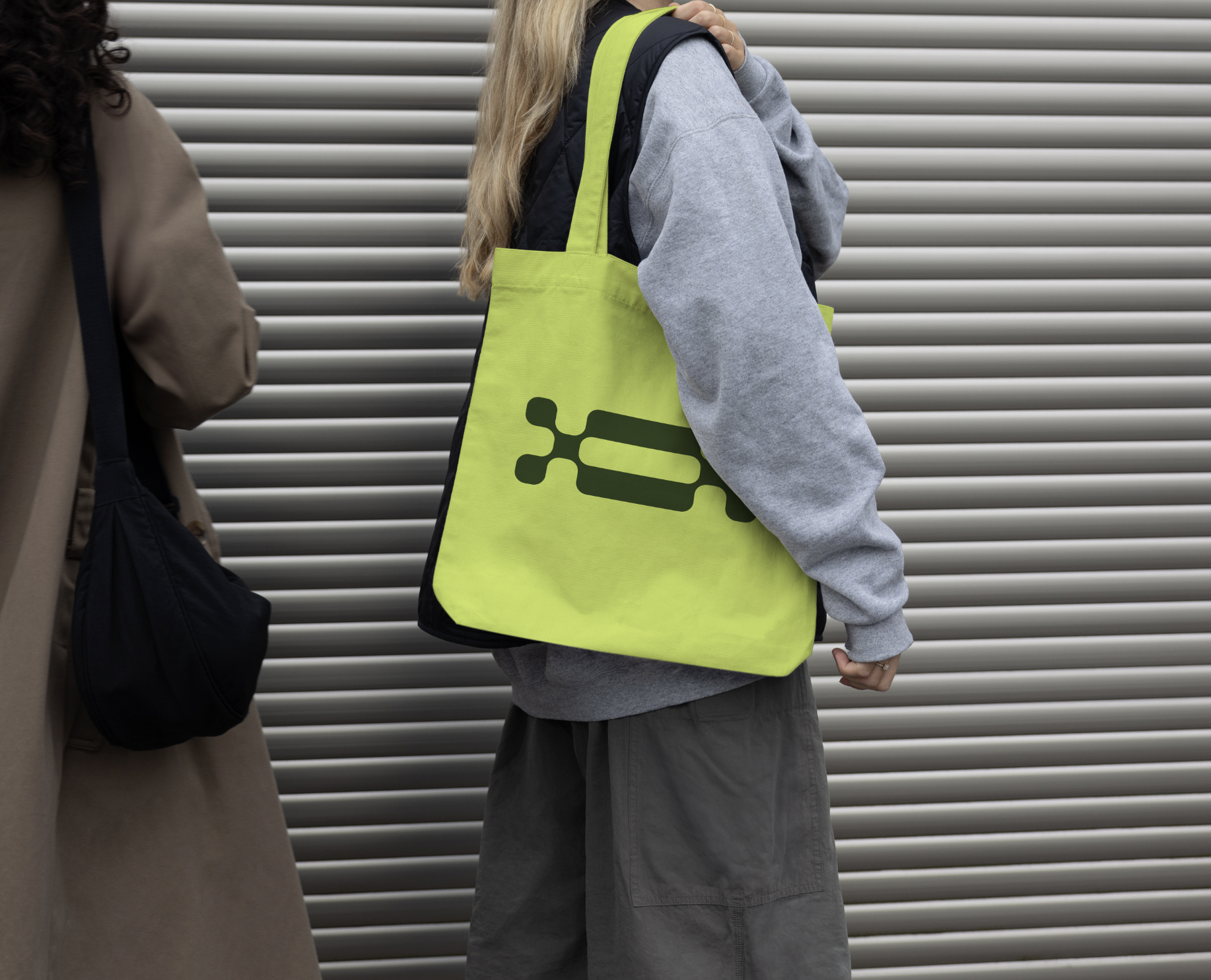Person standing outdoors with a lime green tote bag with the FLEXi logomark, wearing gray hoodie and cargo shorts, against a gray metal slatted background.