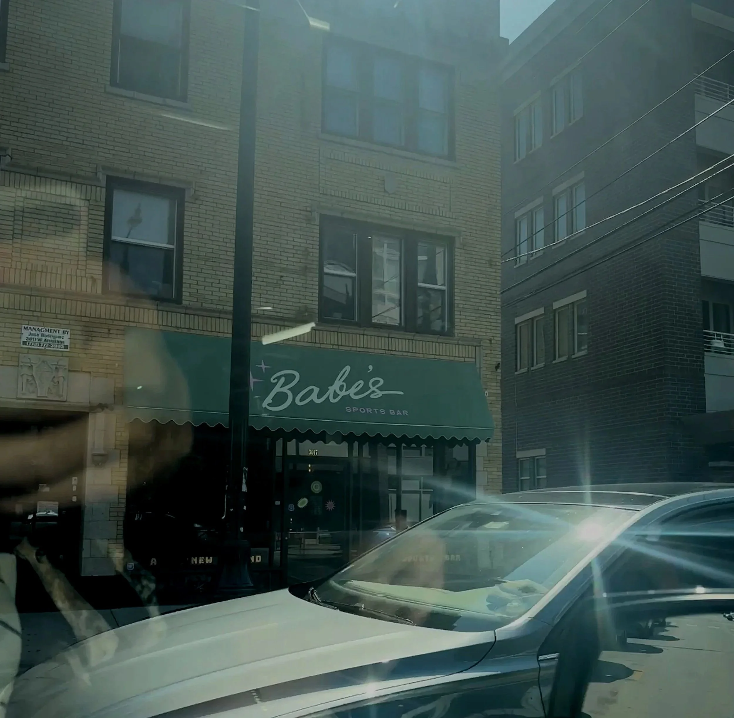 Street view showing a brick building with a green sign that reads 'Babe's Sports Bar,' next to parked cars on the street.
