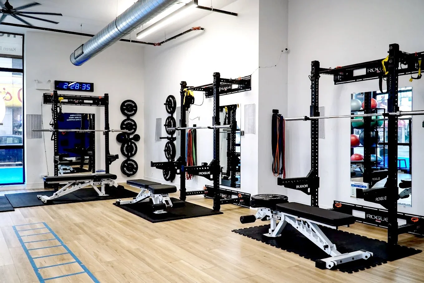 Empty gym with weightlifting equipment including squat racks, benches, weight plates, resistance bands, and medicine balls, with a digital clock on the wall displaying 2:28:35.