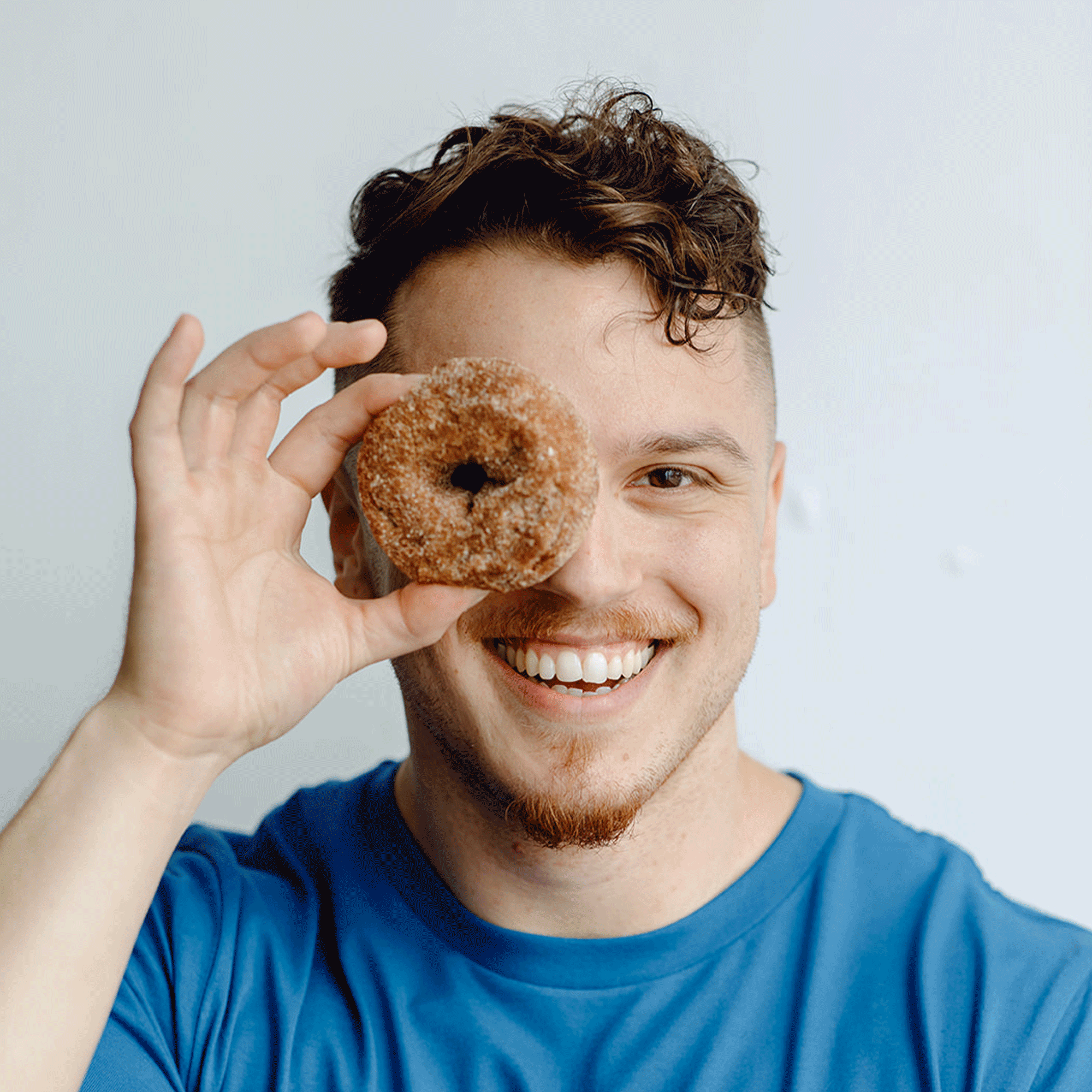 Smiling man in blue shirt holding a cinnamon donut over his right eye.