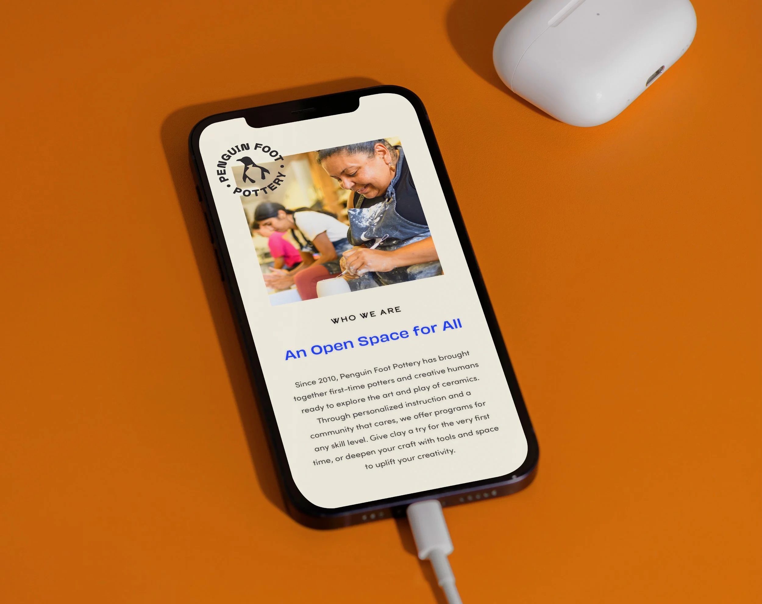 Smartphone on orange surface displaying a webpage for Penguin Foot Pottery with an image of a woman shaping pottery and text describing the community pottery studio.