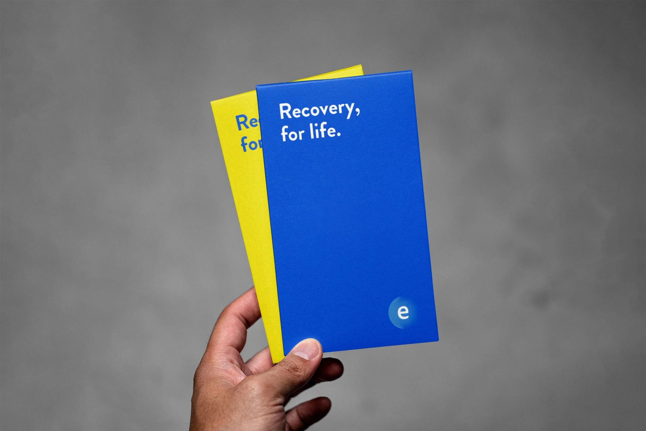 A hand holding two pamphlets, one yellow and one blue, with the blue pamphlet reading "Recovery, for life."