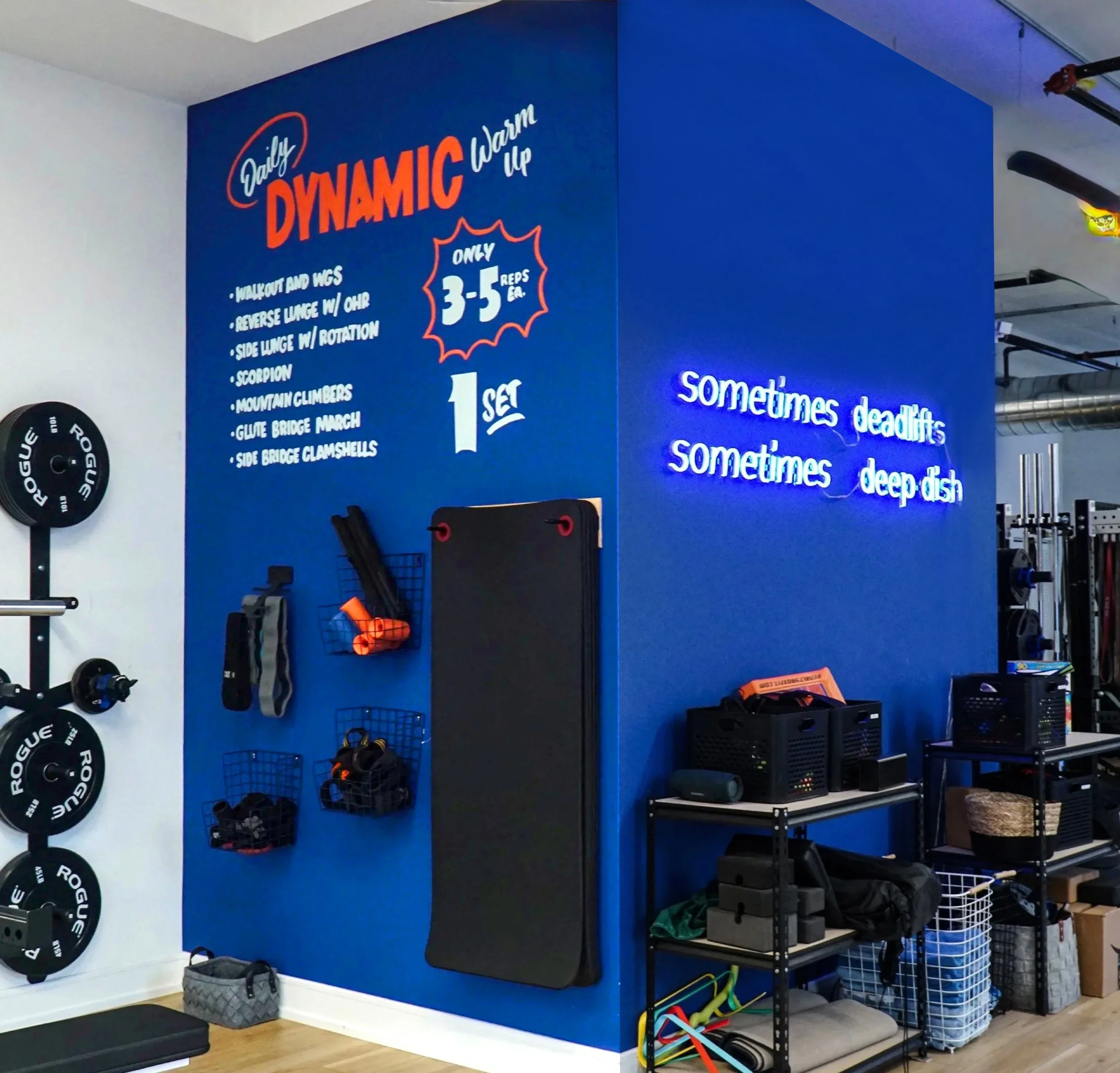 Gym wall with workout instructions and motivational quote in neon lights, weightlifting equipment, and storage shelves.