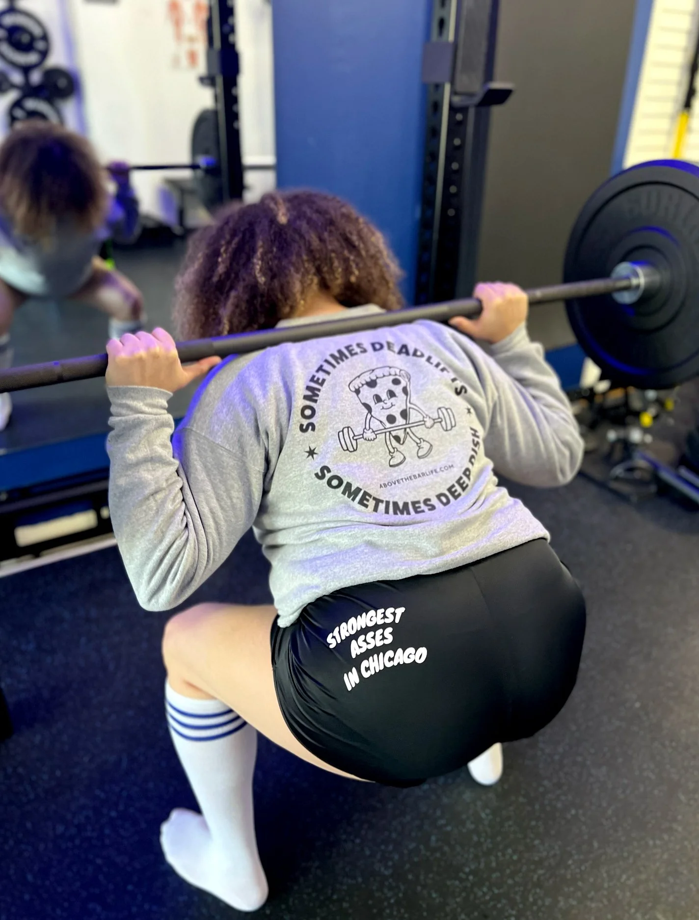 Person in gym squat position with barbell on shoulders, wearing black shorts with writing, striped knee-high socks, and a grey sweatshirt with cartoon character and motivational text. Gym equipment in background.