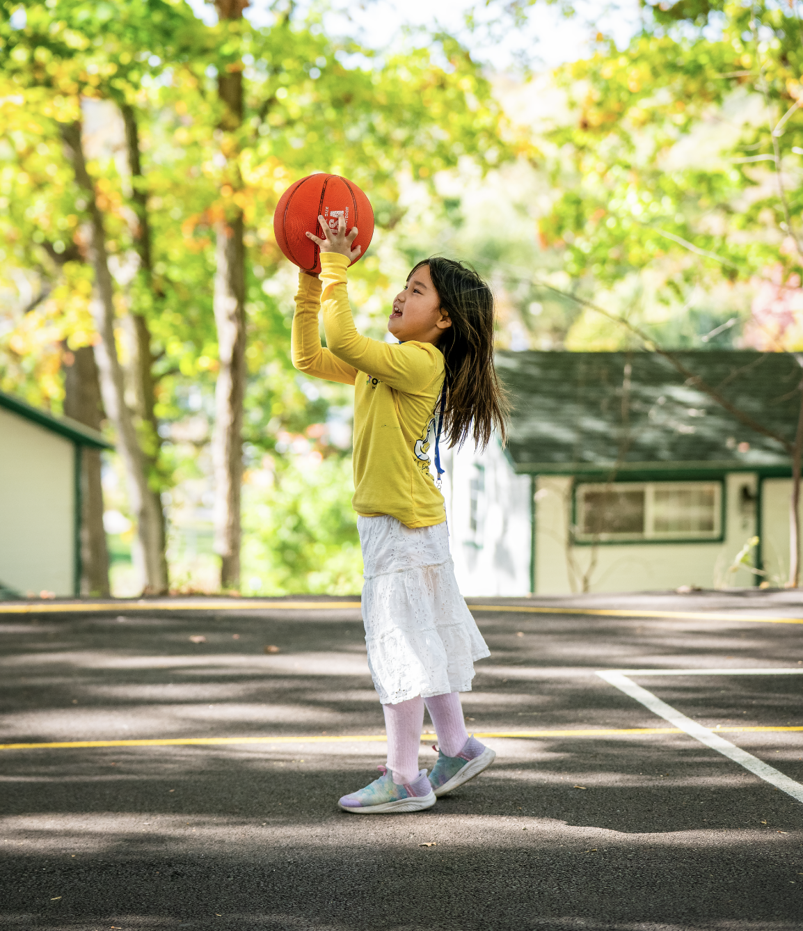 Young girl in a yellow long-sleeve shirt and white skirt playing basketball outdoors on a sunny day.