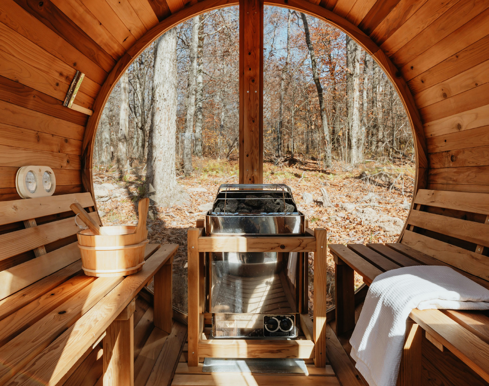 Interior of a wooden sauna with benches, a heater with rocks, and a view of autumn forest outside.