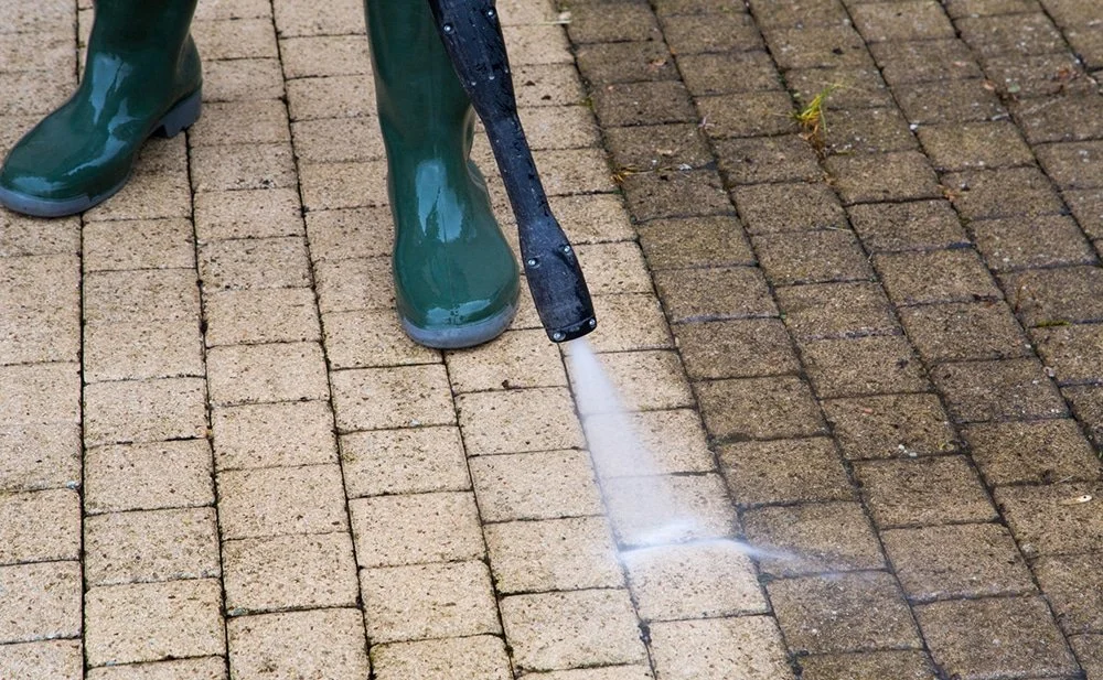 Person in green rain boots pressure washing brick pavement.