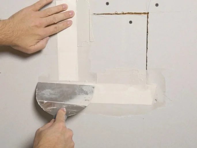 A person holding a drywall taping knife and preparing to tape a seam on a drywall wall.