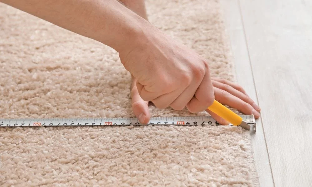 Person measuring a carpet with a tape measure and pencil on a floor.