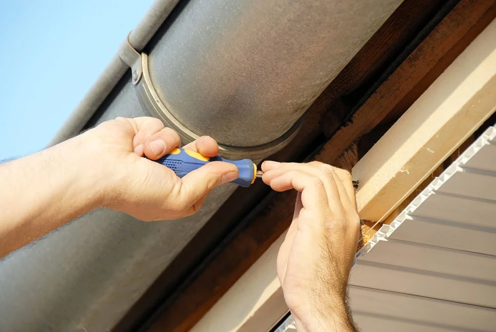 Person using a screwdriver to work on the ceiling near a vent or duct, with visible wood framing and a window blind.