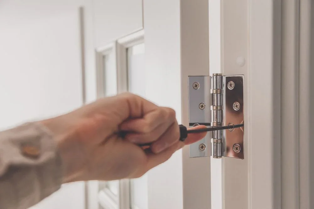 A person using a screwdriver to install or repair a door hinge on a white door.