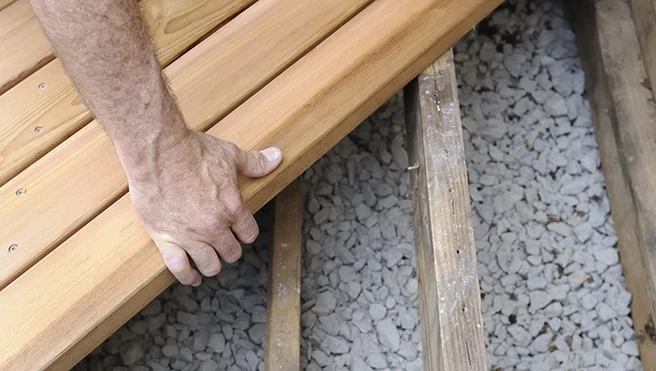 A person is working on a wooden deck, holding a piece of wood in their hand, with gravel below.