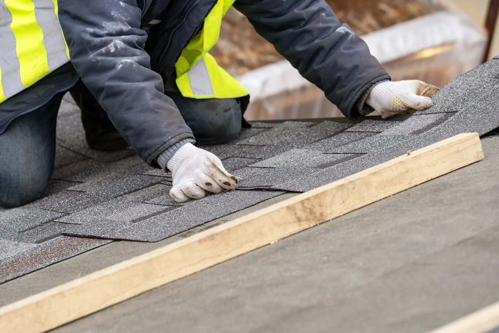 A worker wearing gloves, a high-visibility safety vest, and dark clothing installing asphalt shingles on a roof, kneeling on the shingles with a piece of wood and a construction level nearby.