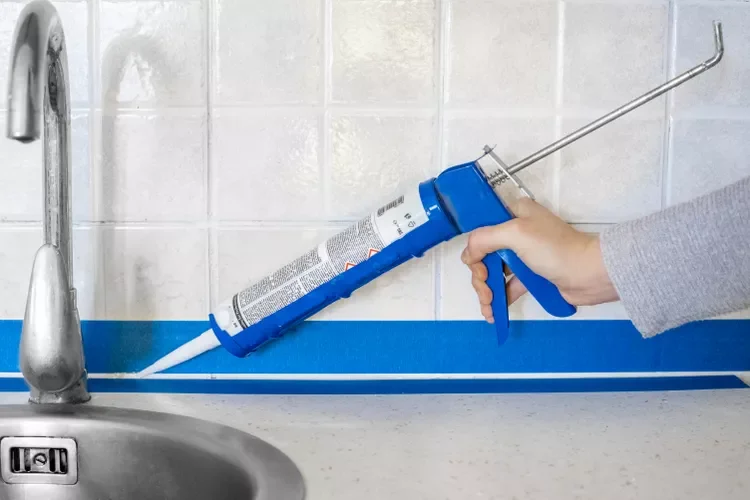 Person using a caulking gun to apply sealant along the edge of a kitchen sink.