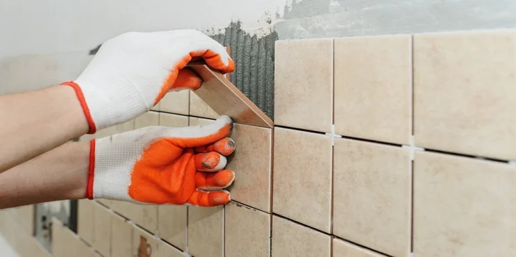 Two hands wearing gloves installing ceramic tiles on a wall with a trowel.