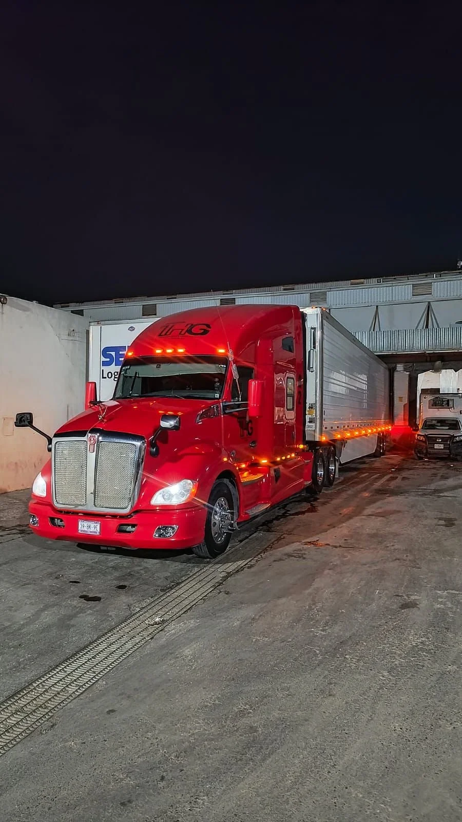 Camión rojo de carga en un área de estacionamiento durante la noche, con otros vehículos y un cielo oscuro en el fondo.