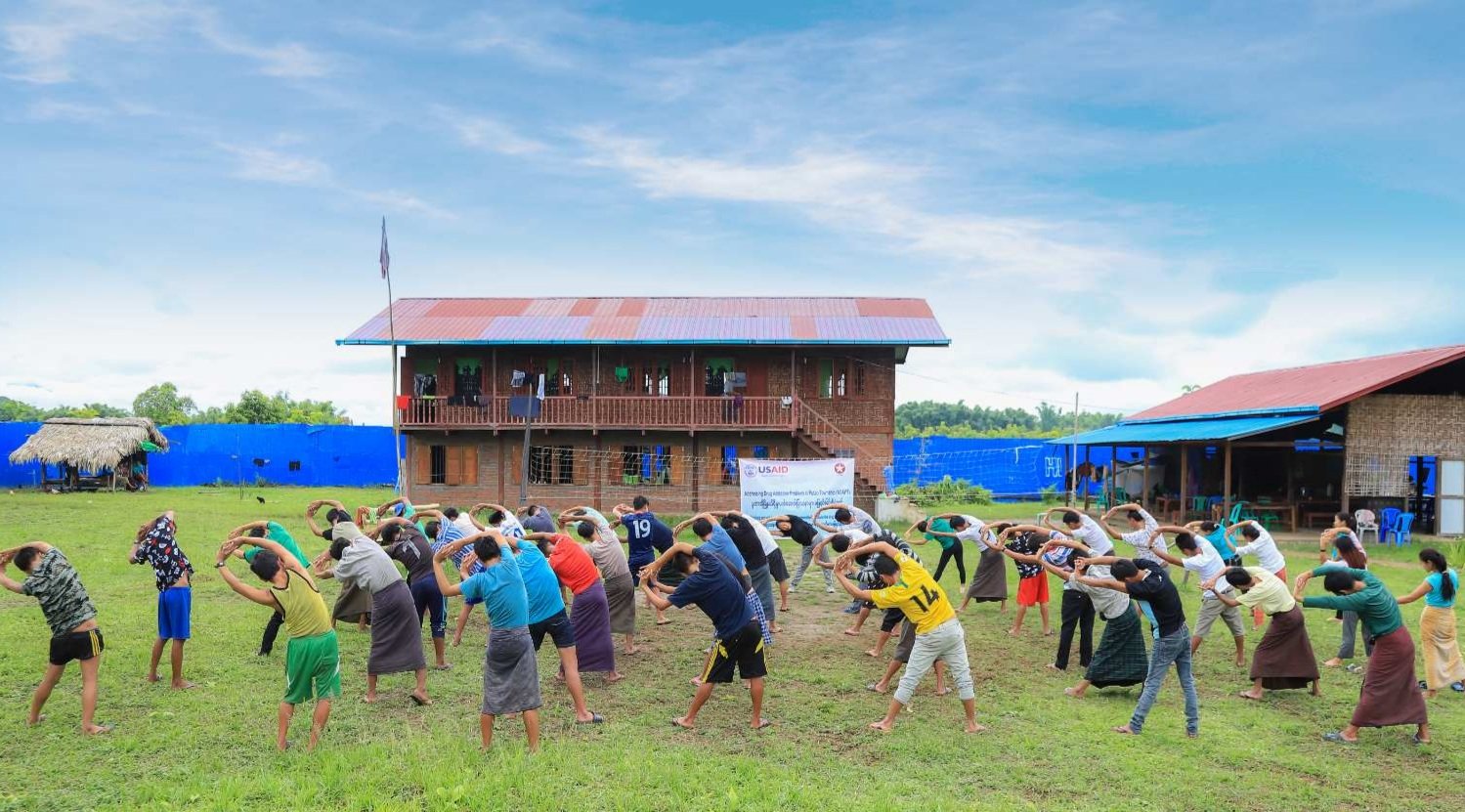 A group of diverse people engaging in outdoor yoga or stretching exercises on a grassy field, with traditional wooden houses and a blue fence in the background under a partly cloudy sky.