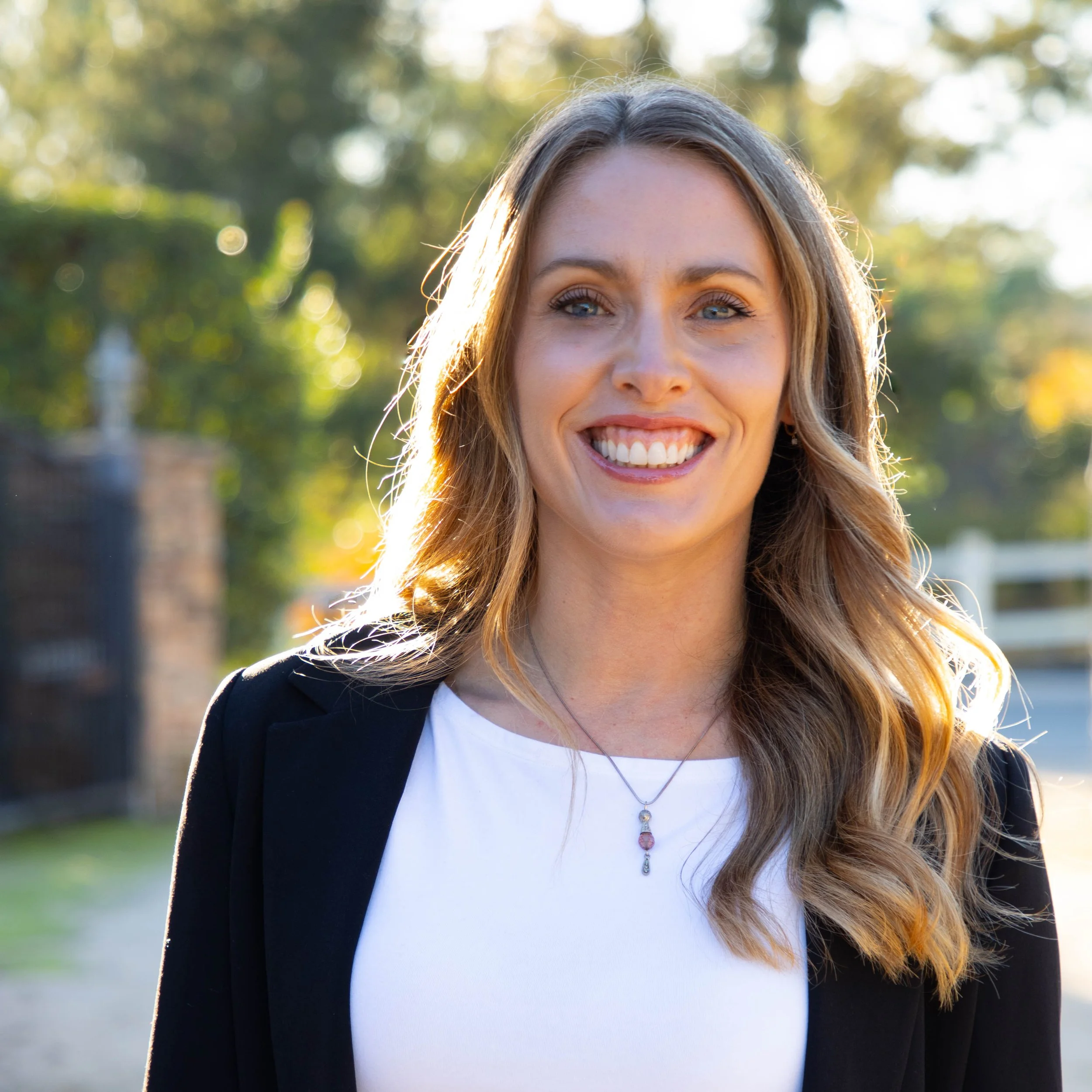 A woman with long wavy blonde hair, smiling outdoors in sunlight, wearing a black blazer over a white top with a necklace. This is Shannon Meehan, Local Works Global co-founder and director of advisory services.