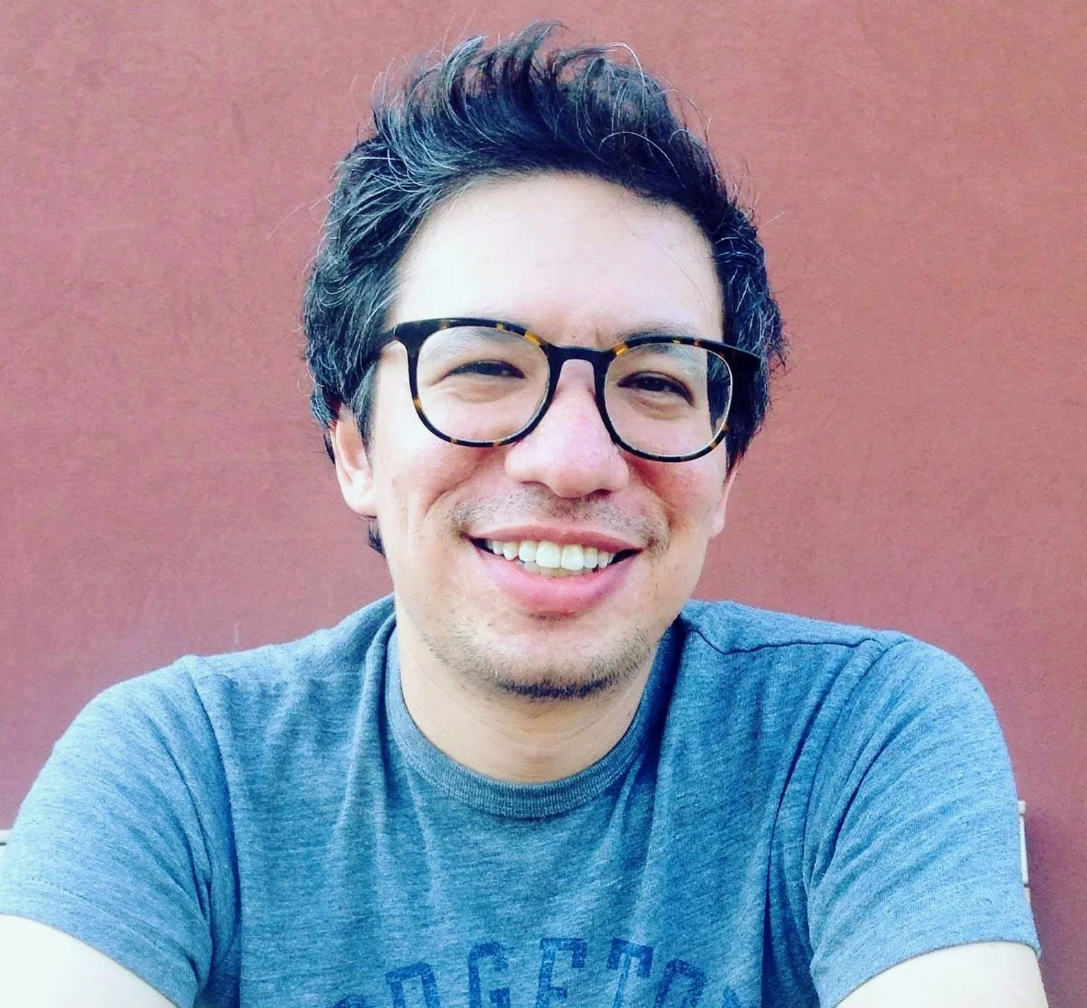 A man with dark, wavy hair and glasses smiling in front of a reddish-brown wall. This is Dan Grant, Local Works Global co-founder and executive director.