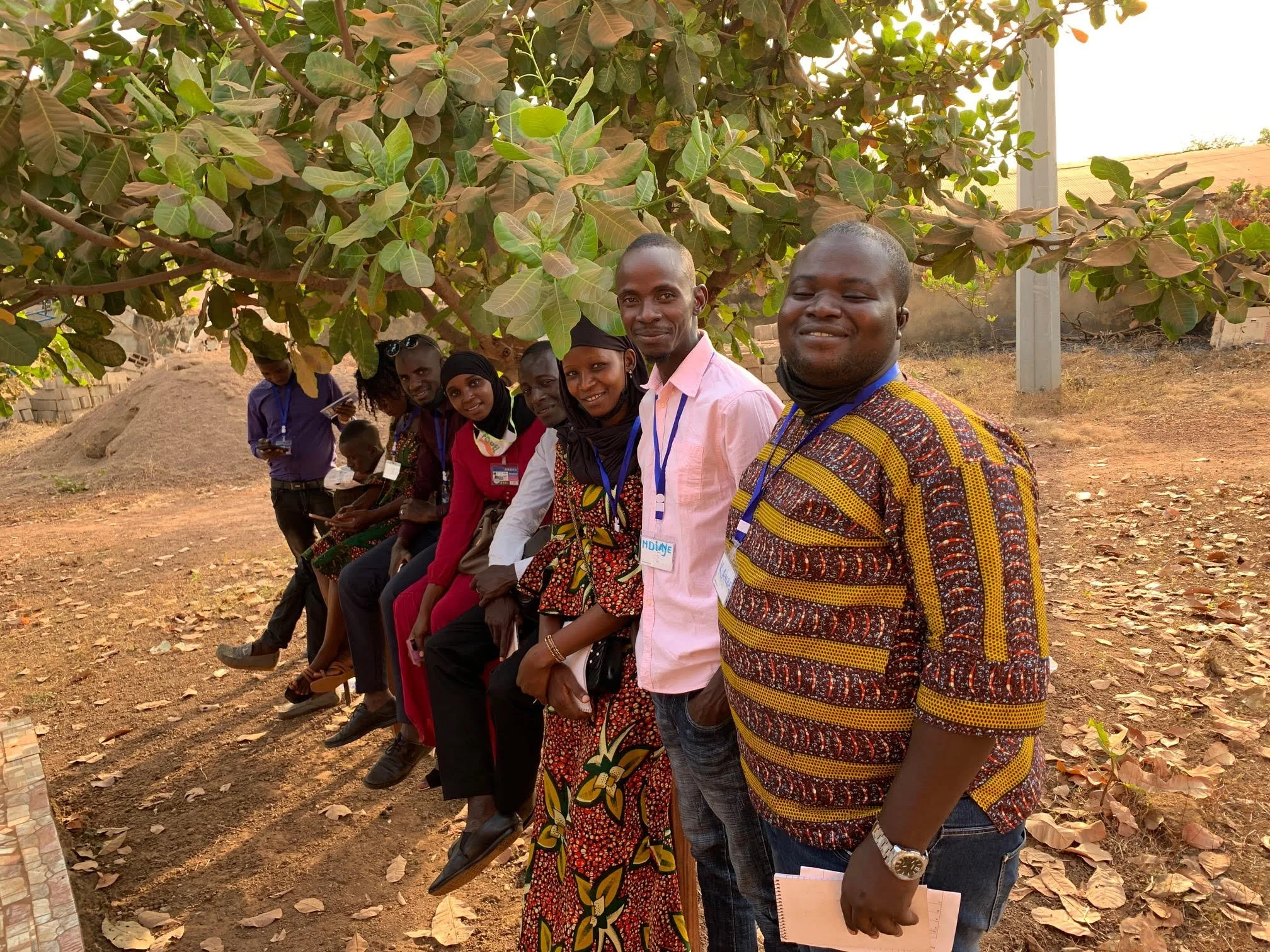 A group of young adults who participated in a youth advisory council in Guinea standing under a large leafy tree outdoors during daytime. They are smiling at the camera, with some holding notebooks and name tags around their necks.