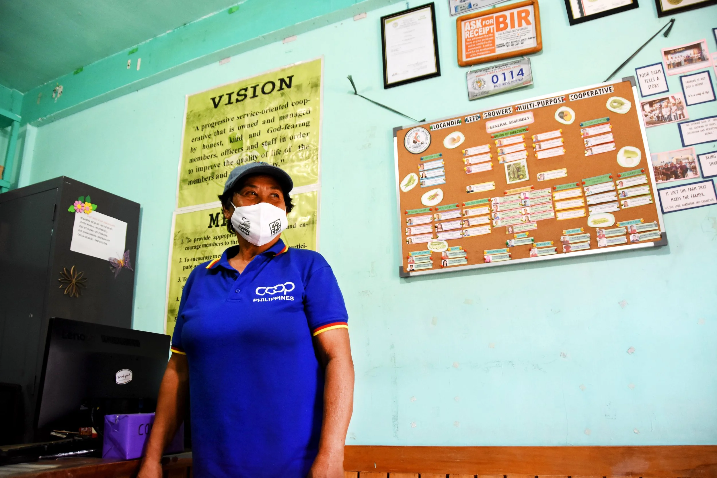 A woman wearing a blue polo shirt with 'Coop Philippines' logo and a face mask, standing in front of a green wall with various posters, including a yellow 'Vision' poster and a bulletin board with photos and notes.