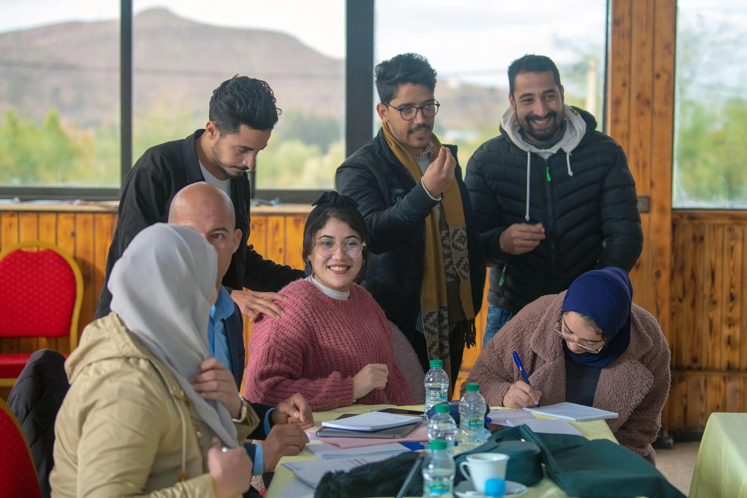 Group of people gathering around a table during a workshop in Morocco in a bright room with wood-paneled walls and large windows.