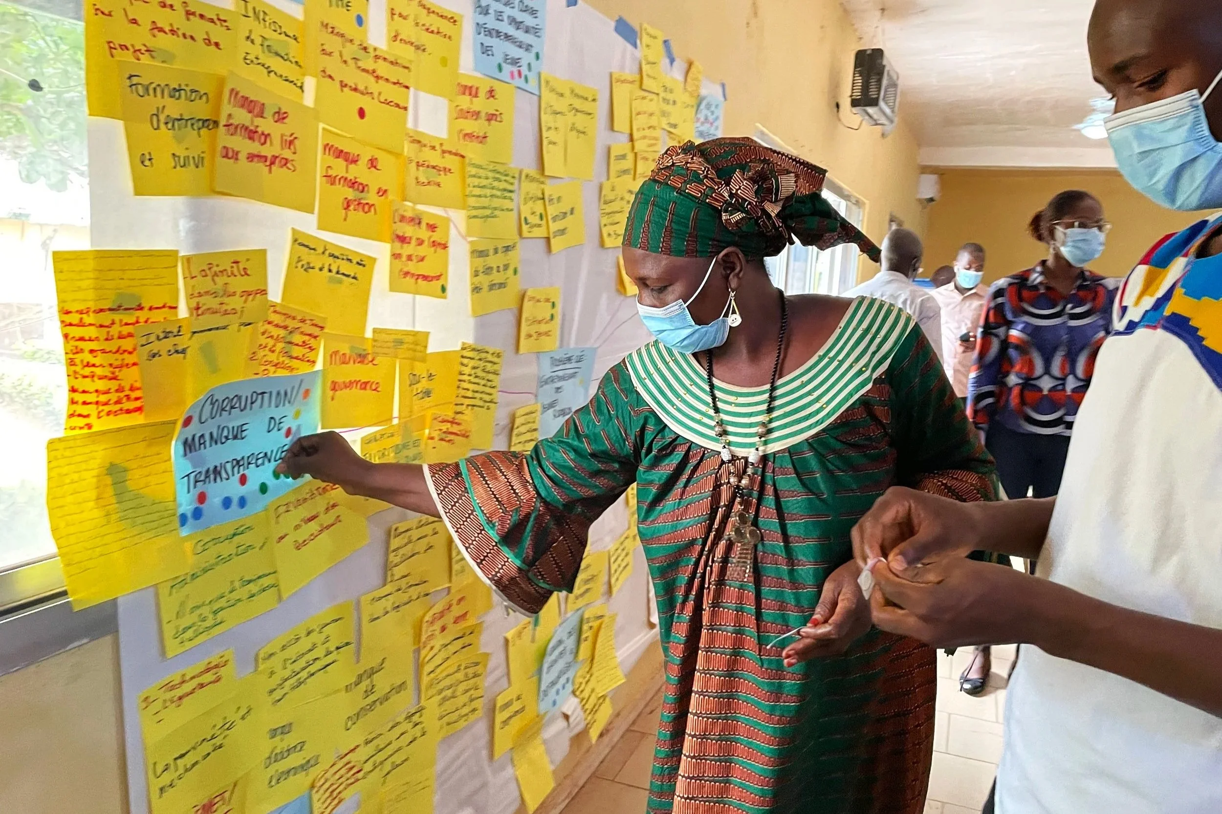 Women wearing face masks examining notes and messages on a wall covered with yellow, blue, and red sticky notes in a room with several other people.