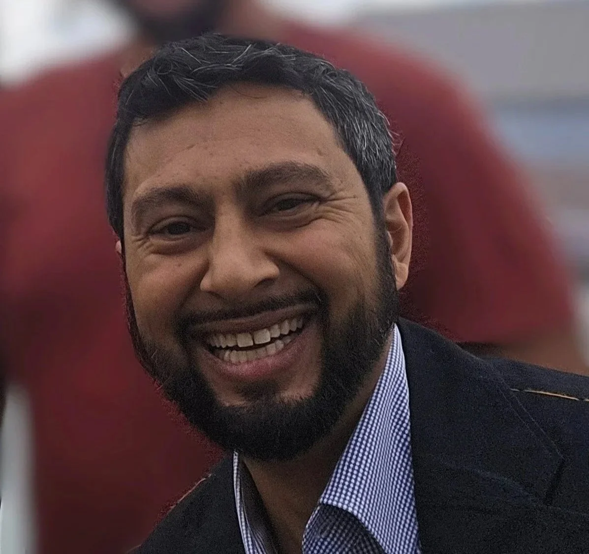 Smiling man with dark hair, beard, wearing a suit jacket and checkered shirt, in front of a blurred background with another person behind him. This is Arjun Tasker, Local Works Global co-founder and senior advisor of partnerships.