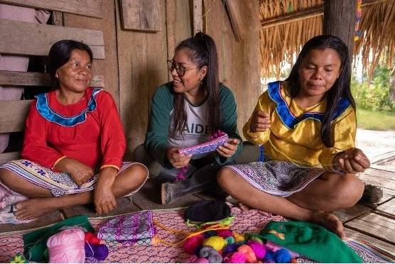 Three women sitting cross-legged inside a wooden hut, engaged in a conversation and sharing colorful woven textiles and yarns.