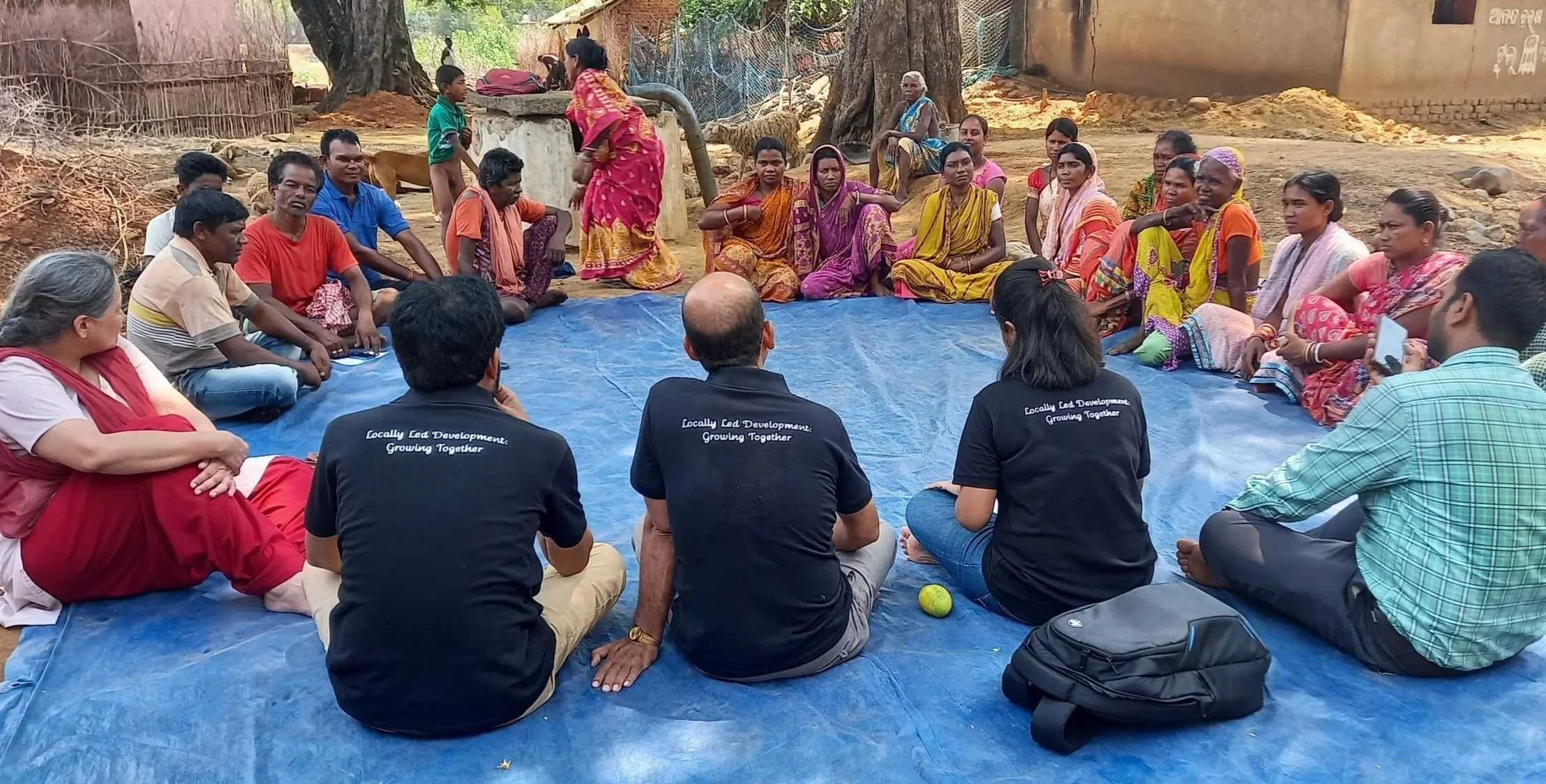 A group of people seated in a circle on a blue tarp outdoors at a listening session in India, with some women wearing traditional colorful saris and some men in casual clothing. In the background, there are trees, a cow, and a few children.