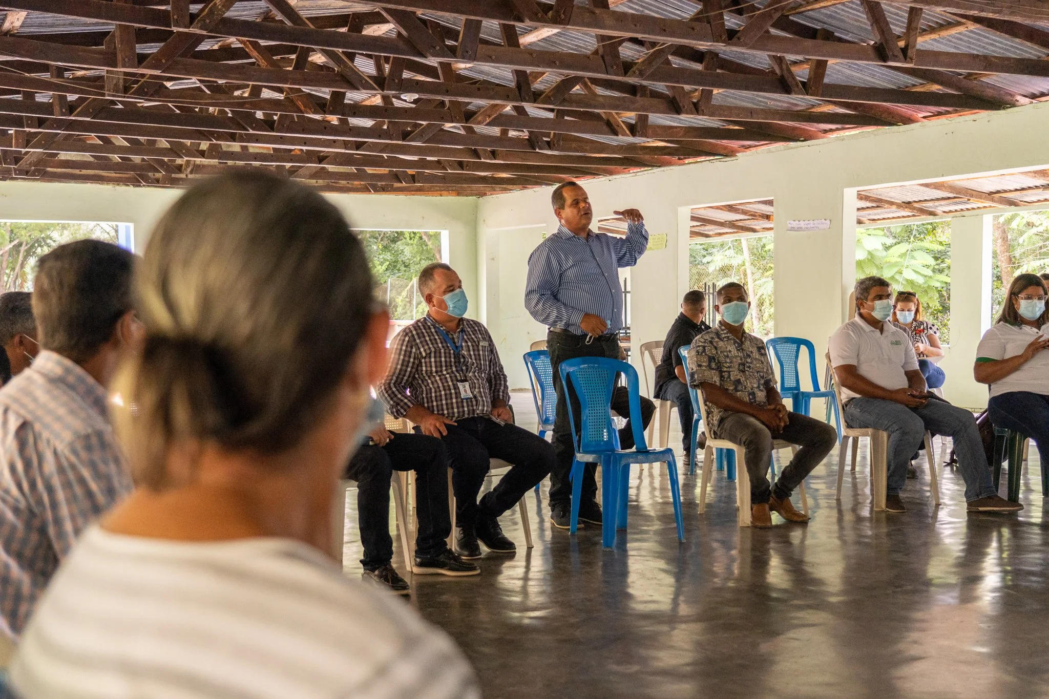 People attending a meeting in a large open room with a high wooden ceiling, some wearing masks, while one man stands and speaks to the group.