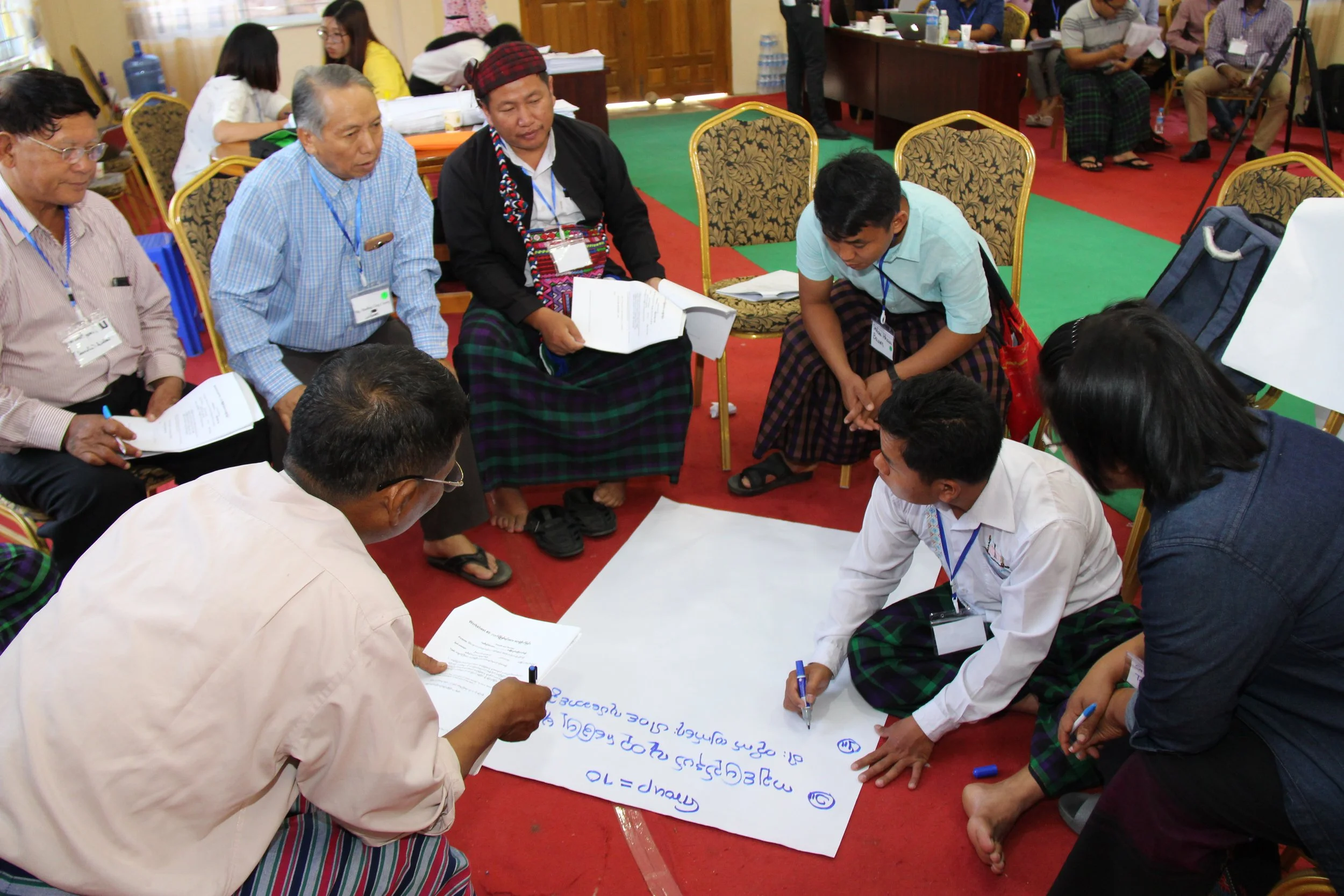A group of people sitting in a circle on the floor, writing on a large sheet of paper during a whole system in the room workshop in Myanmar. Most are wearing name tags and casual attire, with some in traditional clothing.