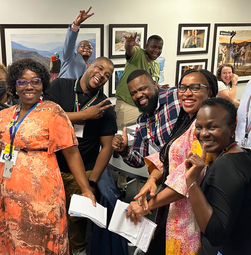 A group of six smiling people at a regional learning summit in South Africa, holding notebooks and making cheerful gestures, with framed artwork on the wall in the background.