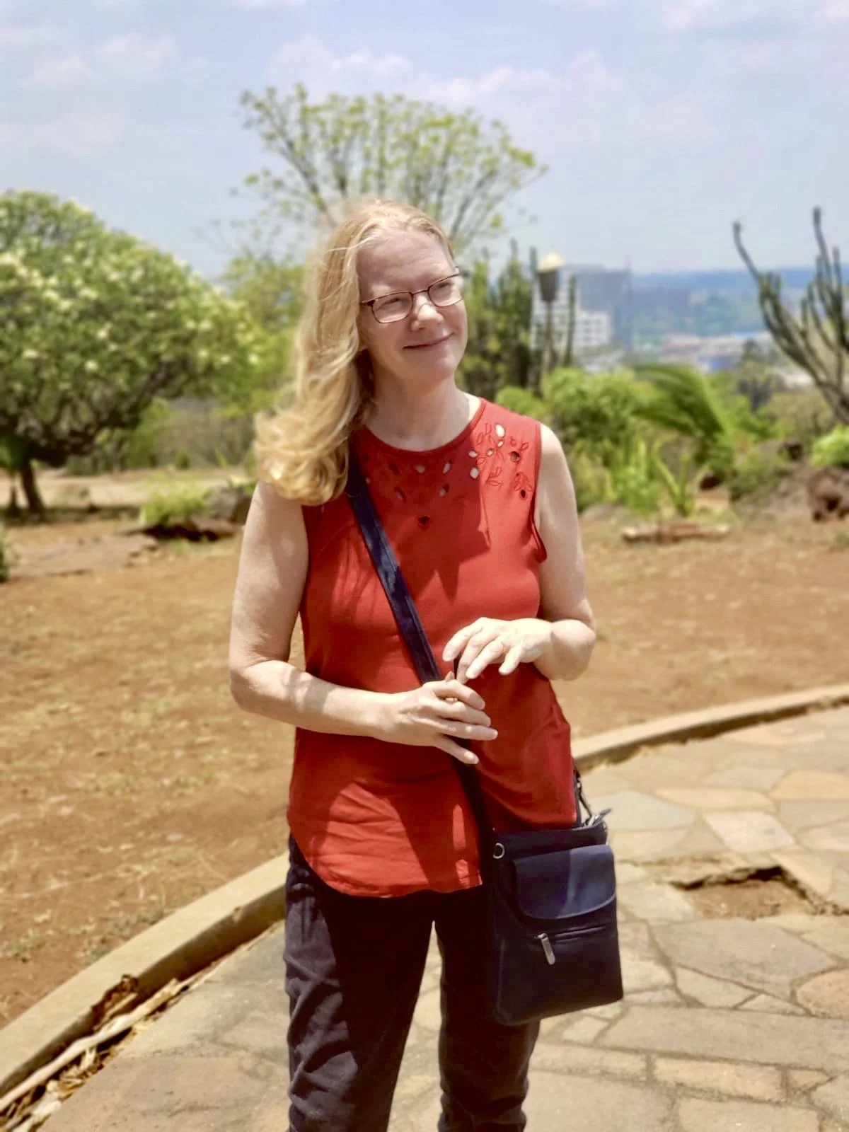 A smiling woman with blonde hair, wearing glasses, a sleeveless red top, and dark pants, standing outdoors on a stone path with greenery and trees in the background. This is Jackie Greene, Local Works Global co-founder and director of strategy.