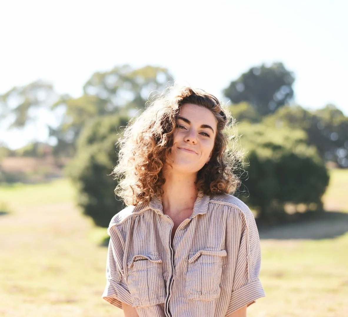 A woman with curly hair smiling outdoors on a sunny day, wearing a striped button-up shirt, with trees and grass in the background. This is Meg Smith, Local Works Global co-founder and director of learning and engagement.