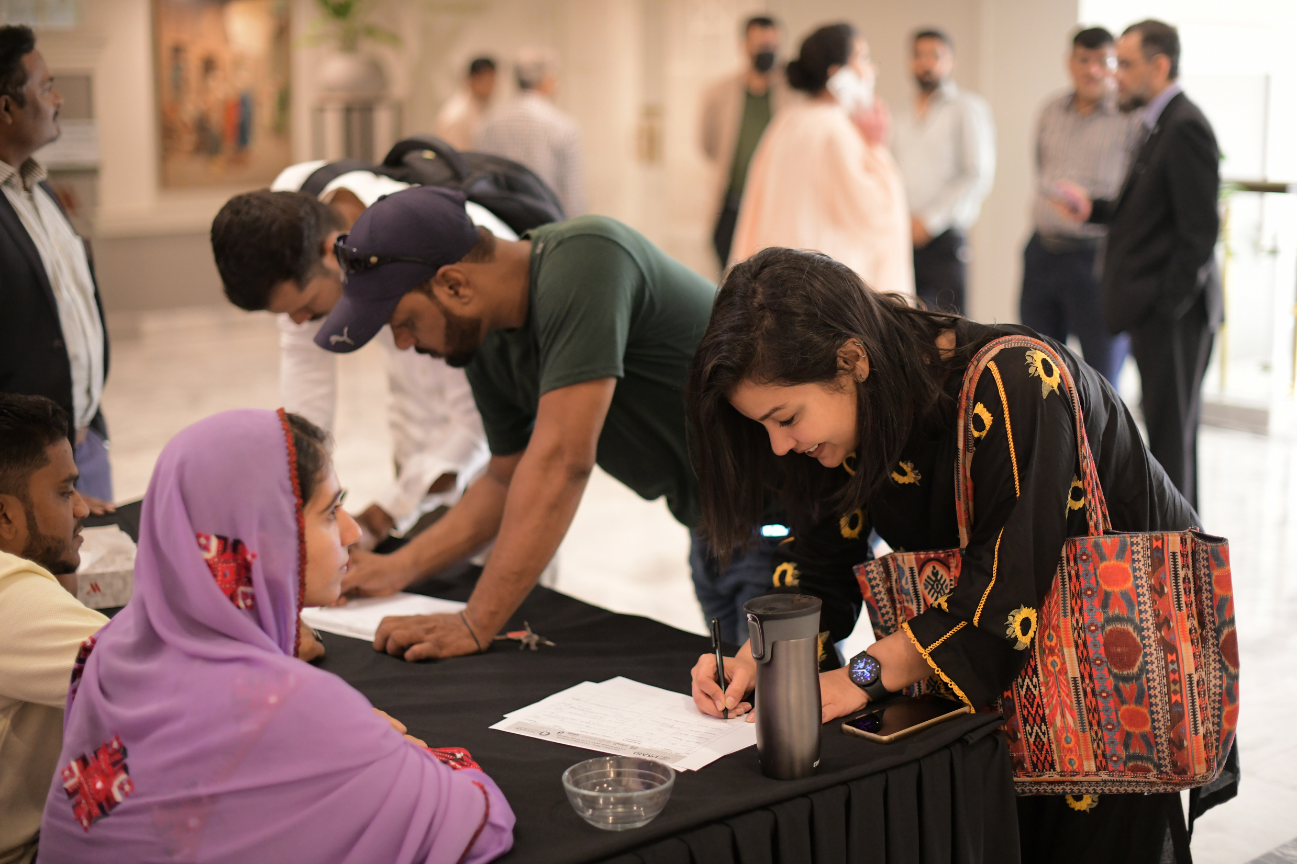 People gather around a registration table at an indoor event in Pakistan, with some filling out paperwork and others waiting or conversing in the background.