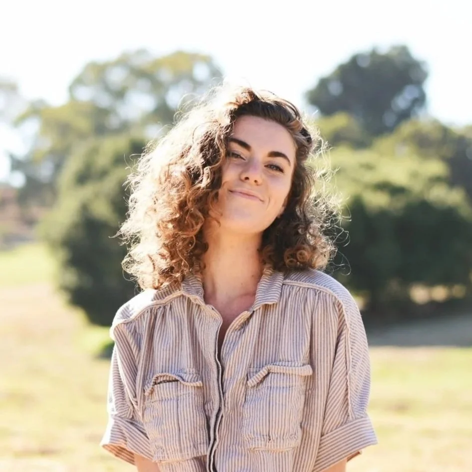 A woman with curly hair smiling outdoors on a sunny day, wearing a striped button-up shirt, with trees and grass in the background. This is Meg Smith, Local Works Global co-founder and director of learning and engagement.